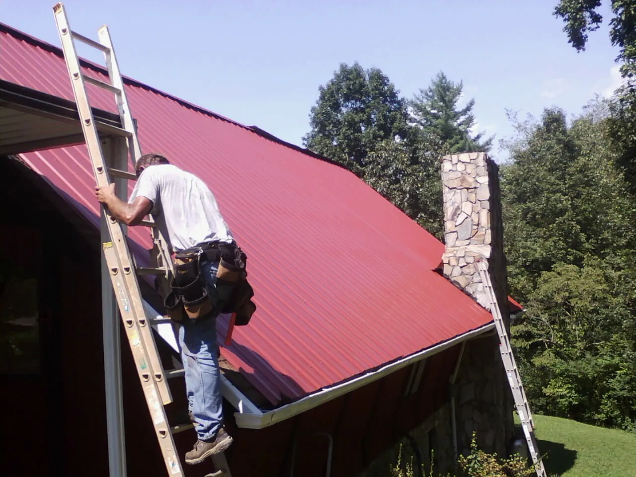 Roofer on a ladder beside a red metal roof and stone chimney — Soffit & Fascia Repair by Roofing Brothers in Martin